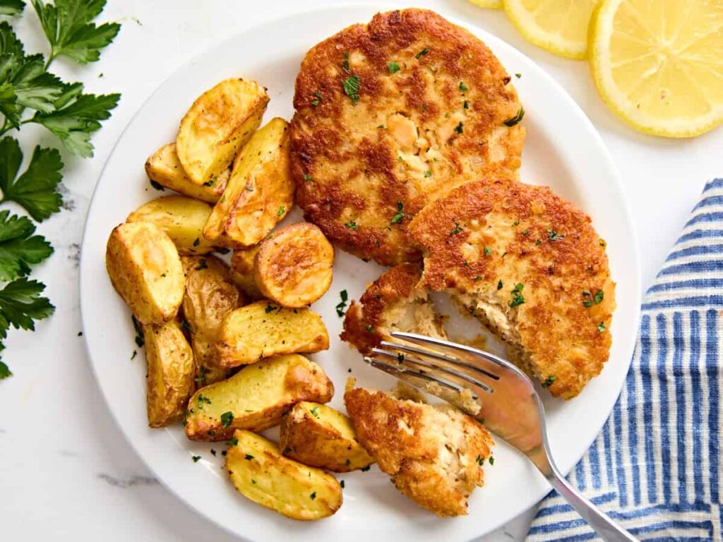 Two salmon patties on a plate, with one sliced in half by a fork, with a side of air fried potatoes.