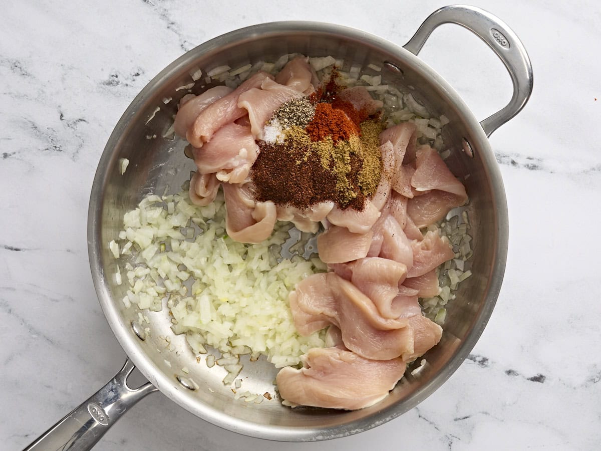 Pechuga de pollo en rodajas y condimentos agregados a las cebollas salteadas y el ajo en una sartén.