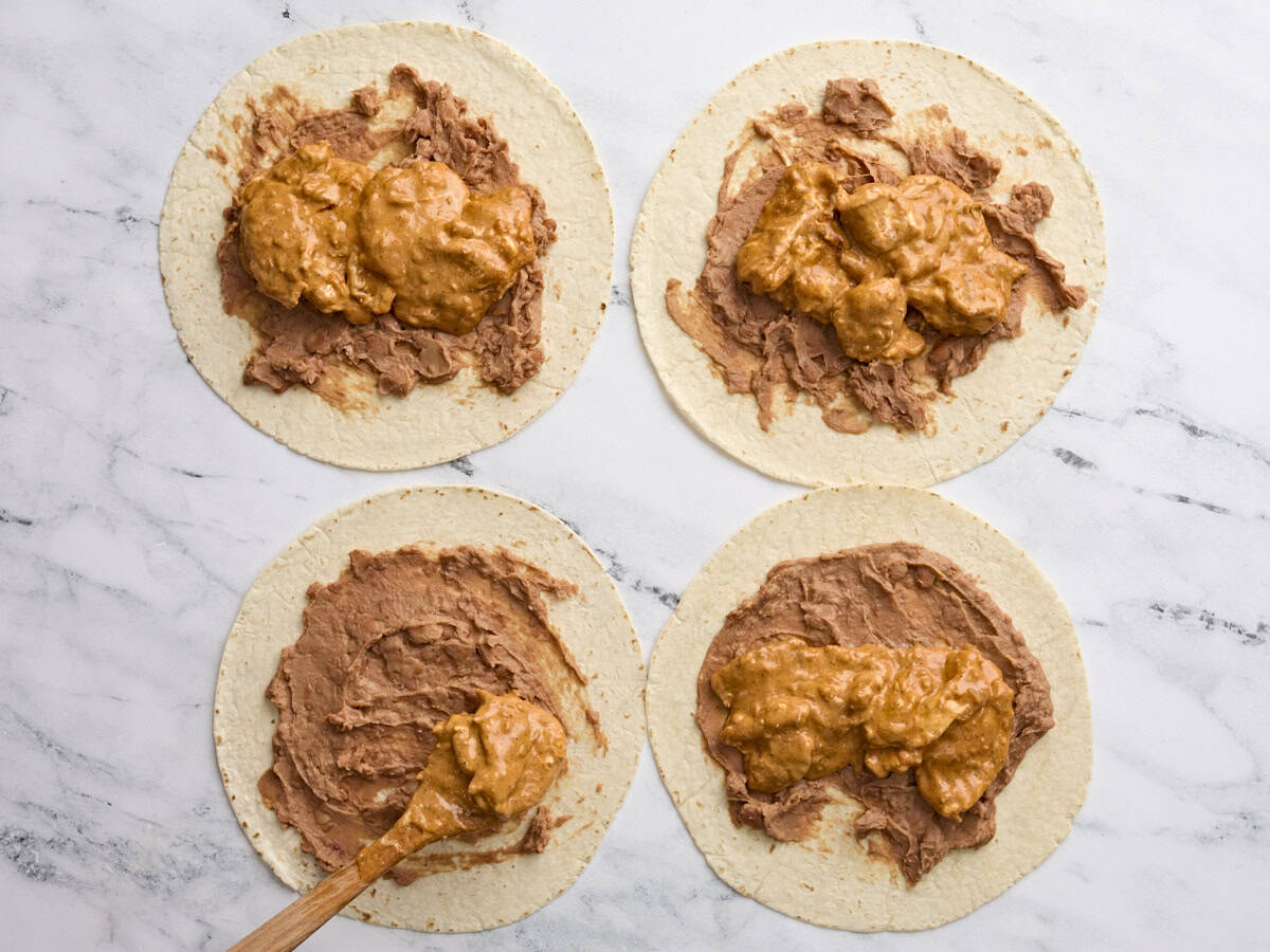 Cuatro tortillas de harina yacen plana sobre una superficie de trabajo, con frijoles refritos extendidos en la parte superior y una cuchara que agrega chimichanga de pollo llenado en la parte superior.