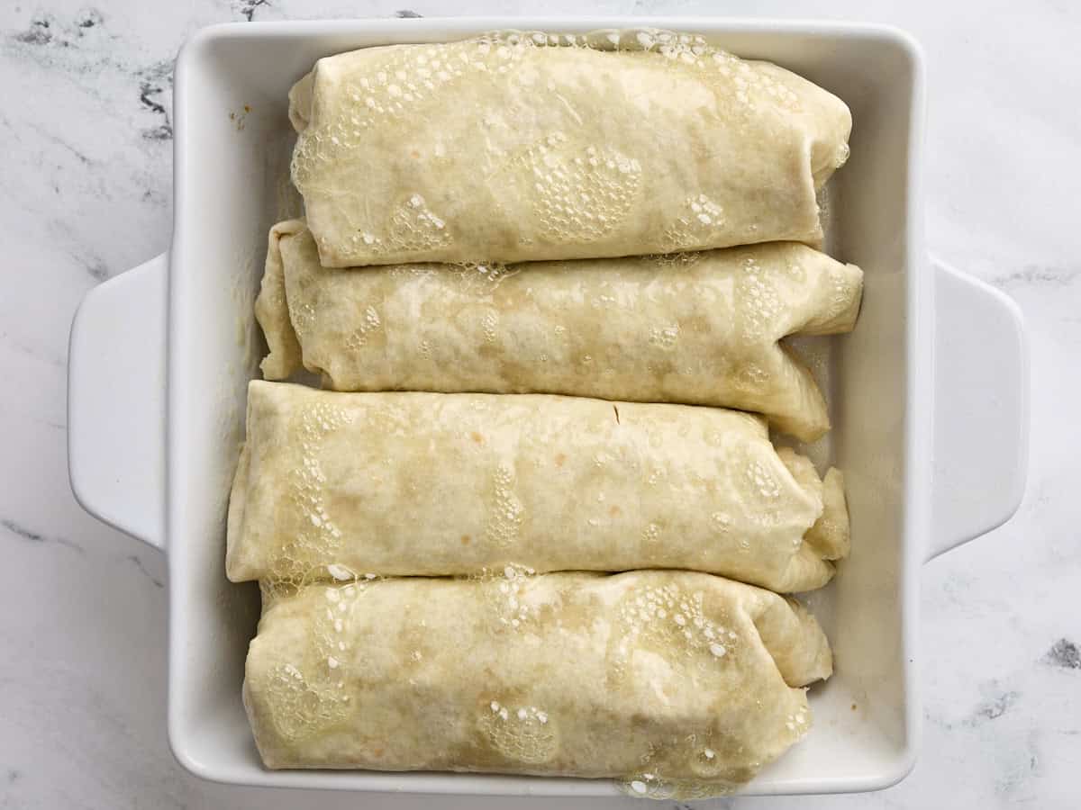 Chimichangas de pollo doblado en una fuente para hornear, cubierta con aerosol de aceite de cocina.