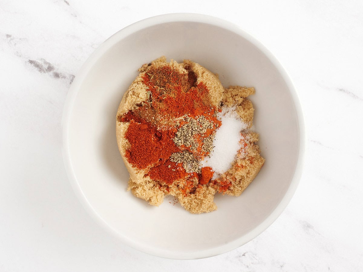 Azúcar morena, cayena, ajo en polvo, pimentón, sal, pimienta negra, clavo, canela y tomillo en un tazón.