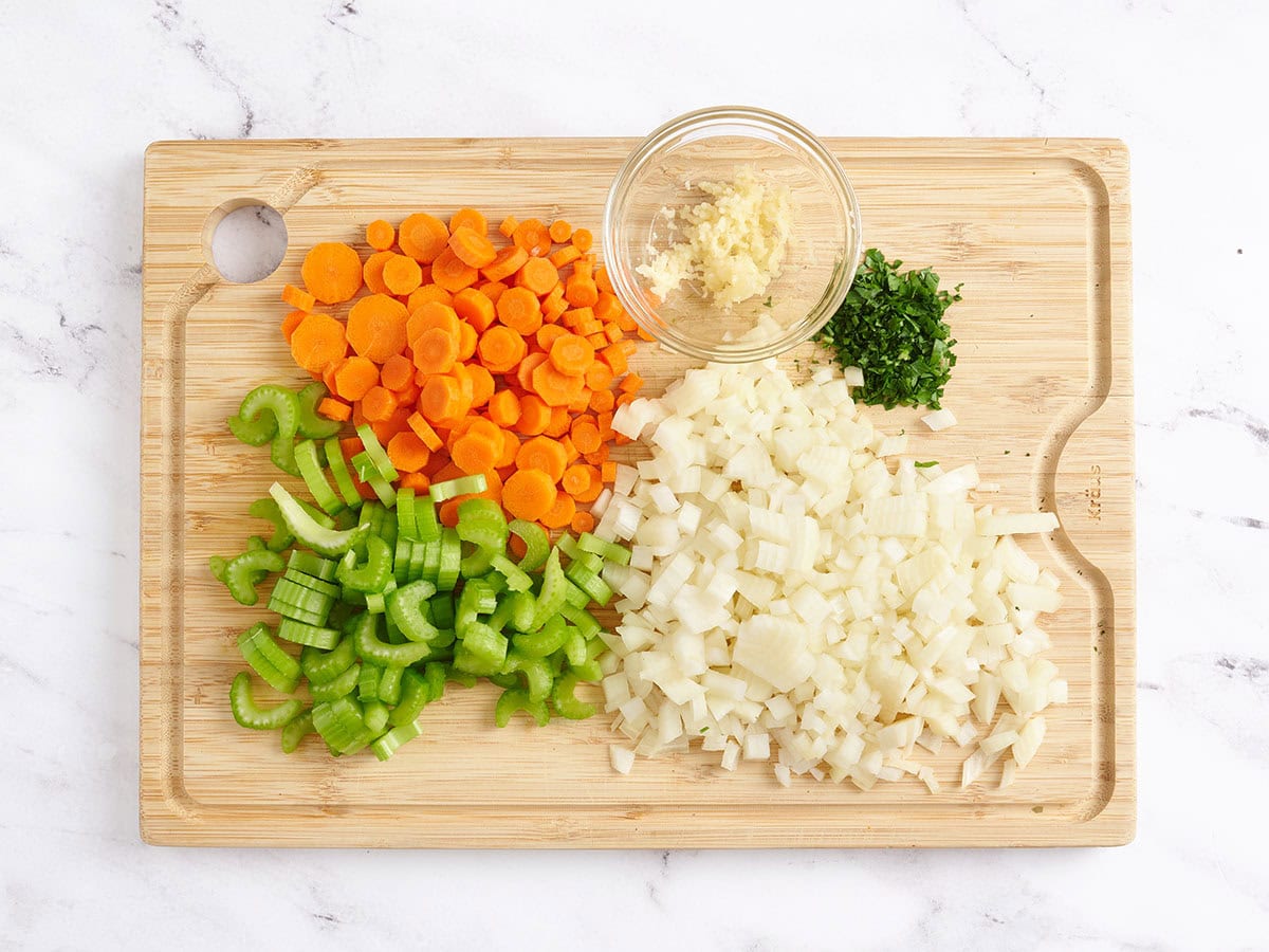 Las zanahorias cortadas en cubitos, el apio, la cebolla, el perejil picado y el ajo picado en una tabla de corte de madera.
