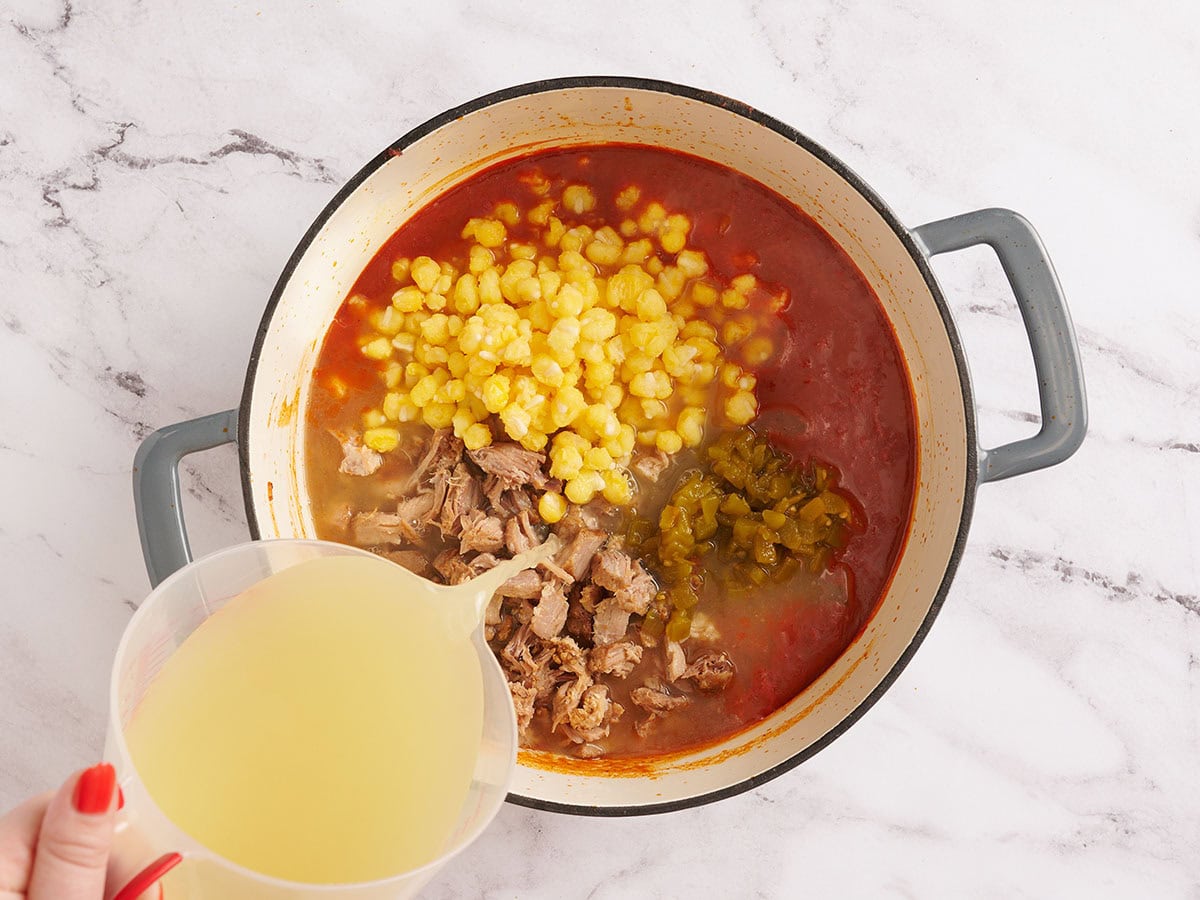 Se agrega carne de cerdo desmenuzada, chile verde, maíz molido y caldo a una olla para hacer posole.