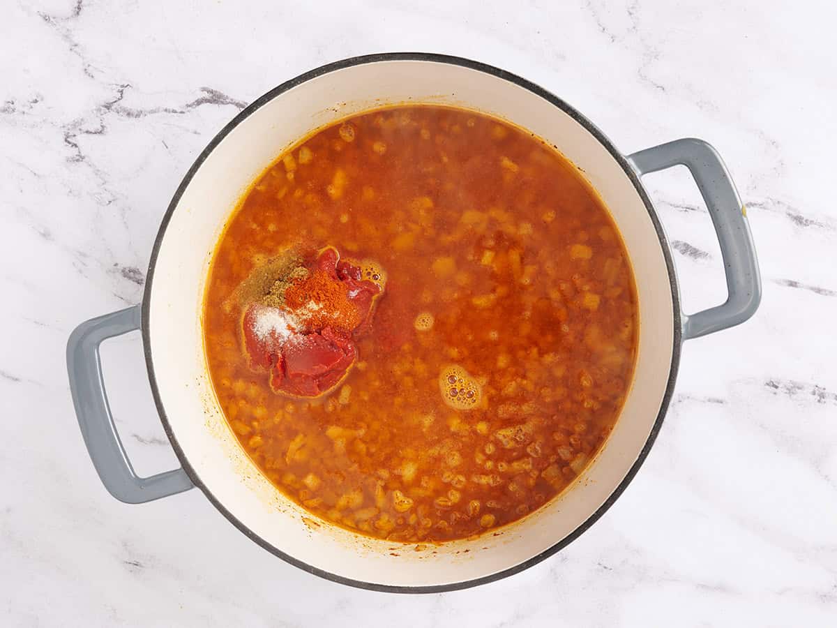 Se agrega pasta de tomate, condimentos y agua a las cebollas salteadas en una olla.