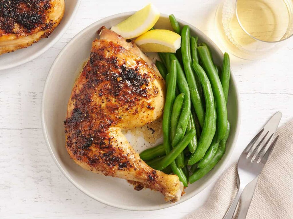 Overhead view of a baked chicken leg on a plate with green beans and lemon wedges.
