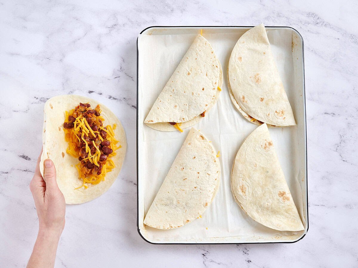 Una mano doblando una quesadilla junto a una bandeja para hornear con otras quesadillas.
