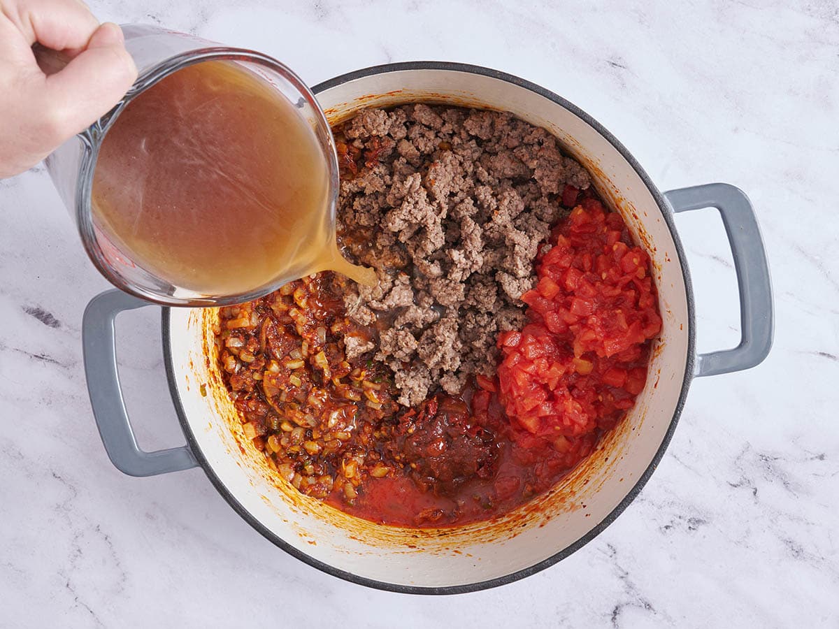 Tomates cortados en cubitos, carne molida dorada y caldo de carne agregados a una olla de cebollas salteadas.