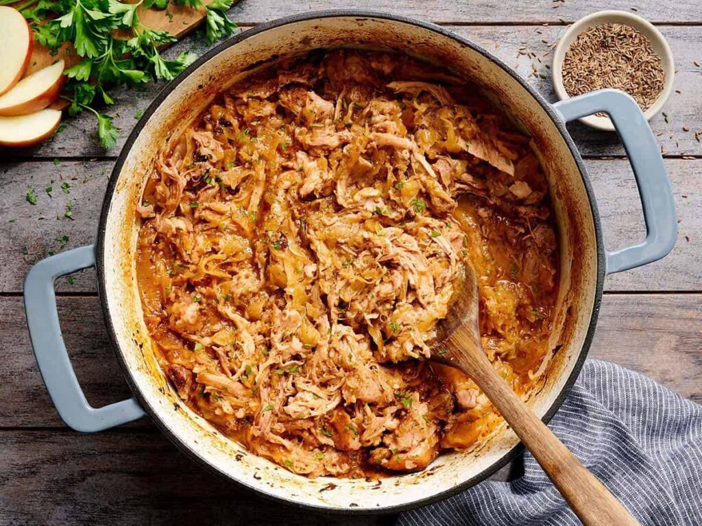 Overhead view of shredded pork and sauerkraut in a pot with a wooden spoon.