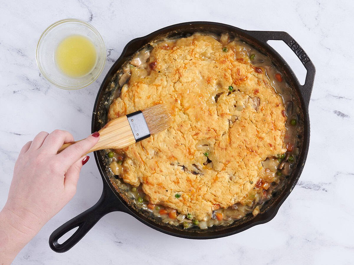 Una mano cepillando mantequilla derretida sobre un pastel de pollo.