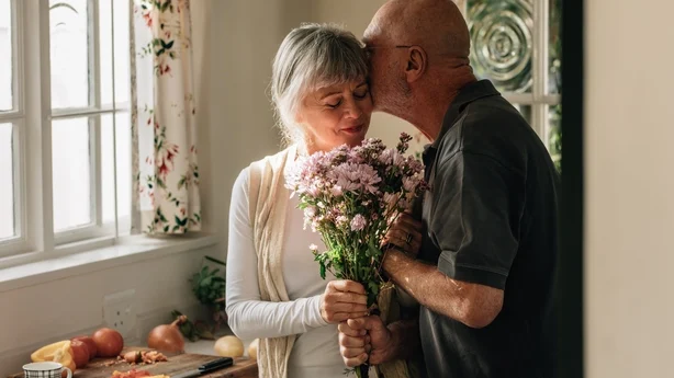 Una pareja mayor de pie en la cocina sosteniendo un ramo de flores