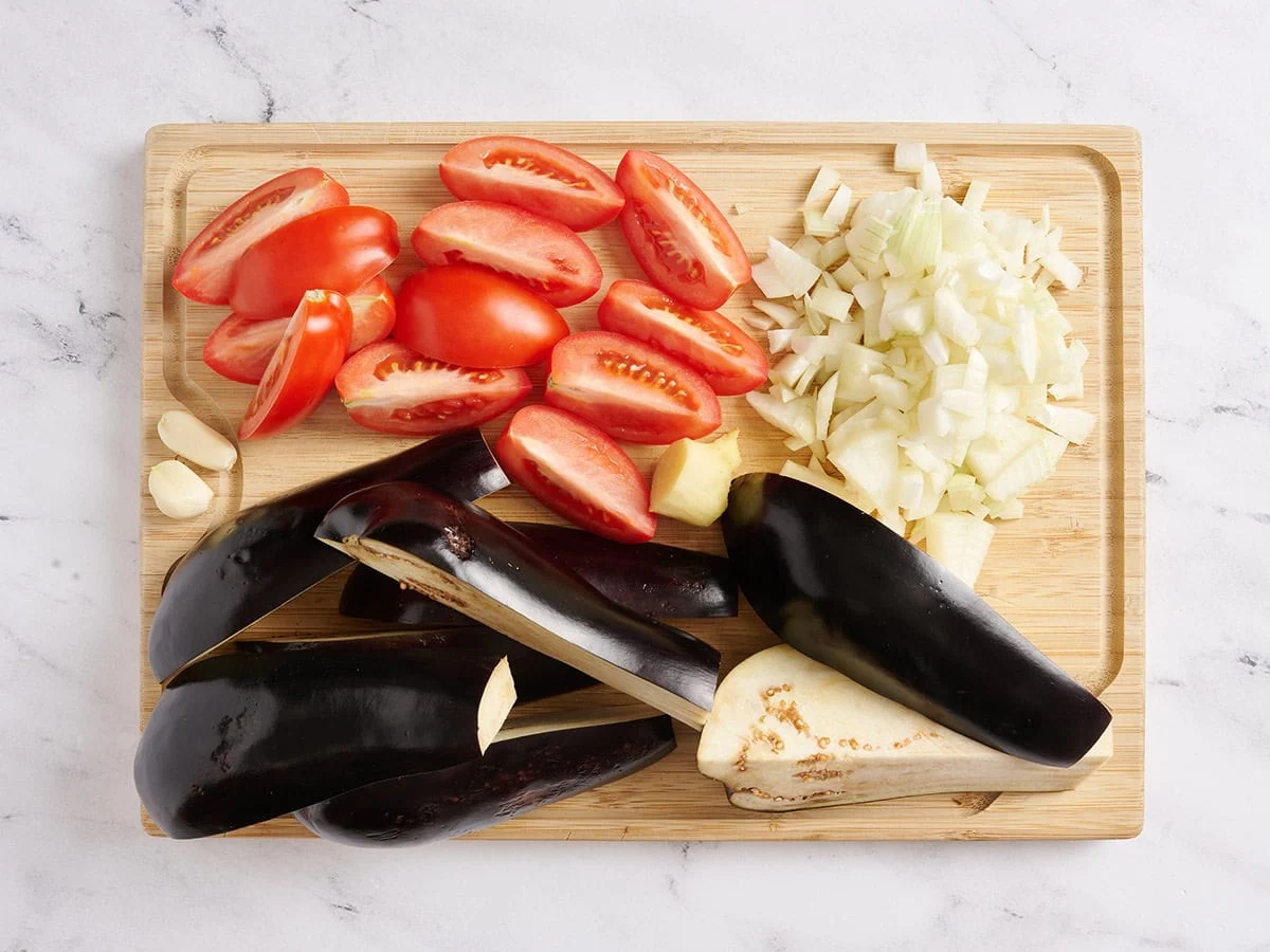Tomates y berenjenas en cuartos, cebolla picada y dientes de ajo pelados sobre una tabla de cortar de madera.