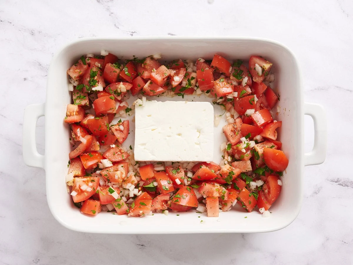 Tomates y cebollas cortados en cubitos en una fuente para horno con un bloque de queso feta en el medio.