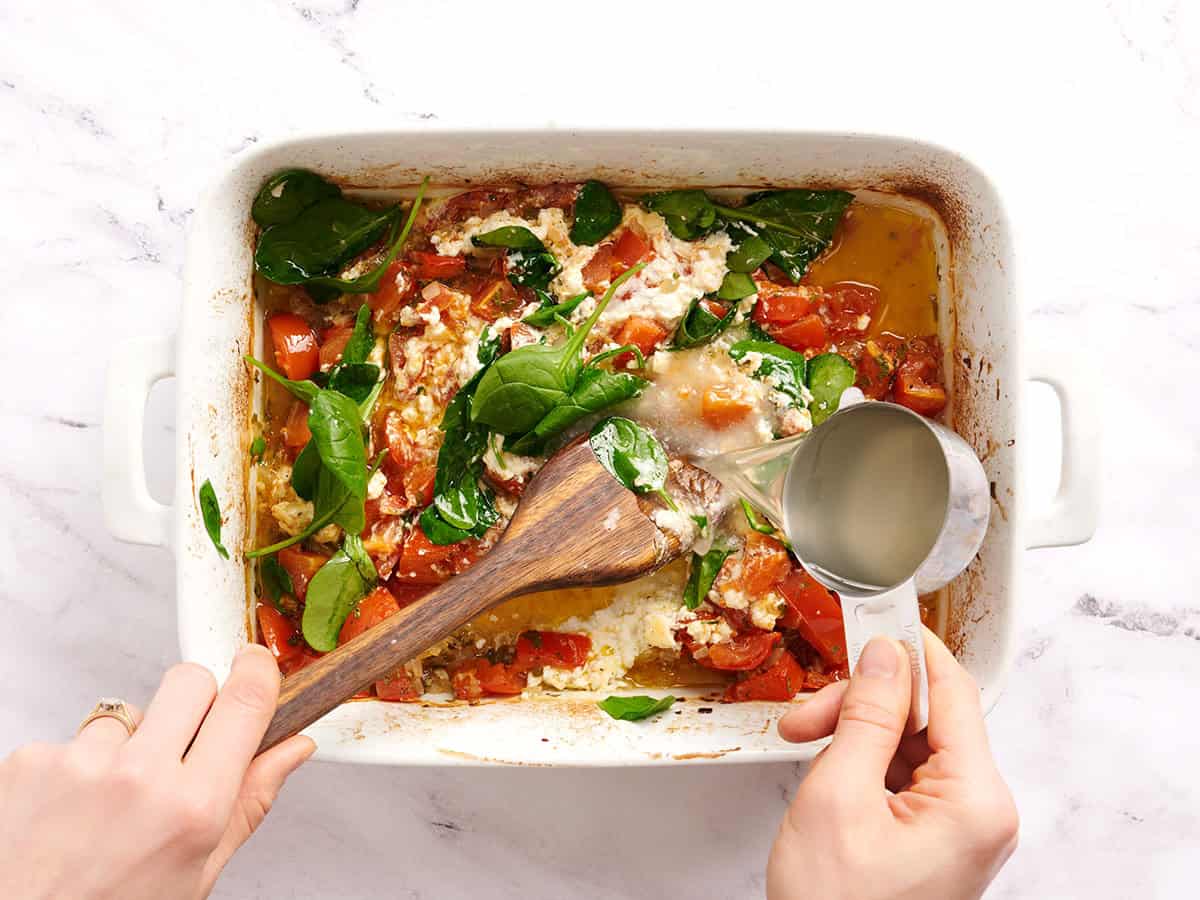 Una mano vierte el agua de pasta reservada en una fuente para hornear con tomates horneados y queso feta.