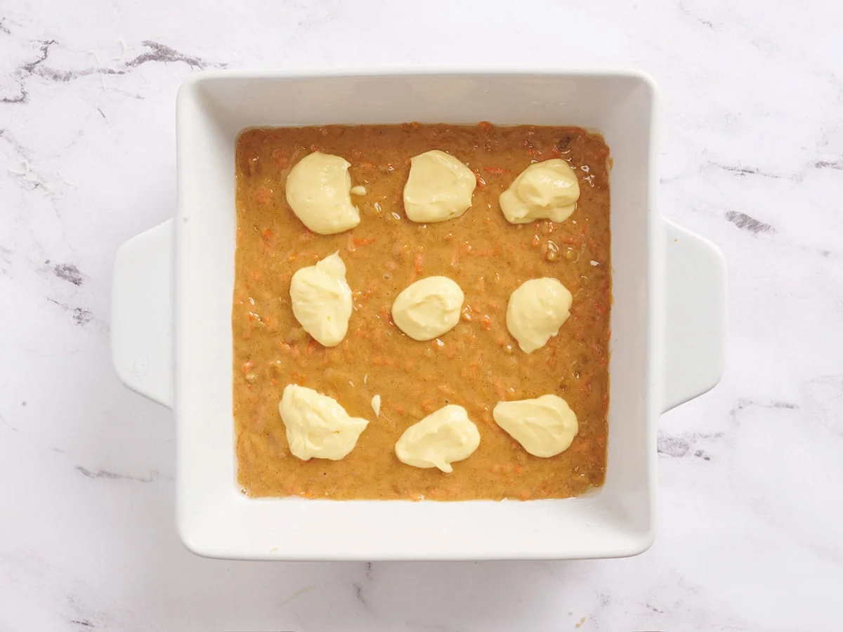 Se agrega una capa de mezcla de barra de pastel de zanahoria a una fuente para hornear con la mezcla de pastel de queso encima.