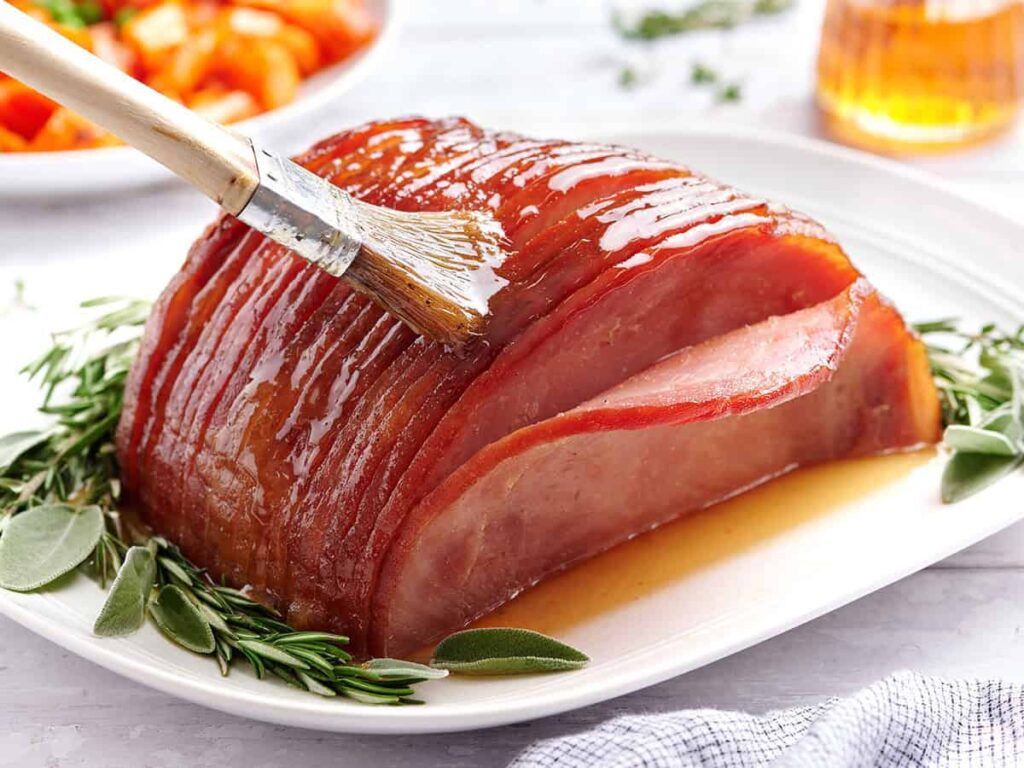Honey glazed ham on a plate with herbs, being brushed with glaze.