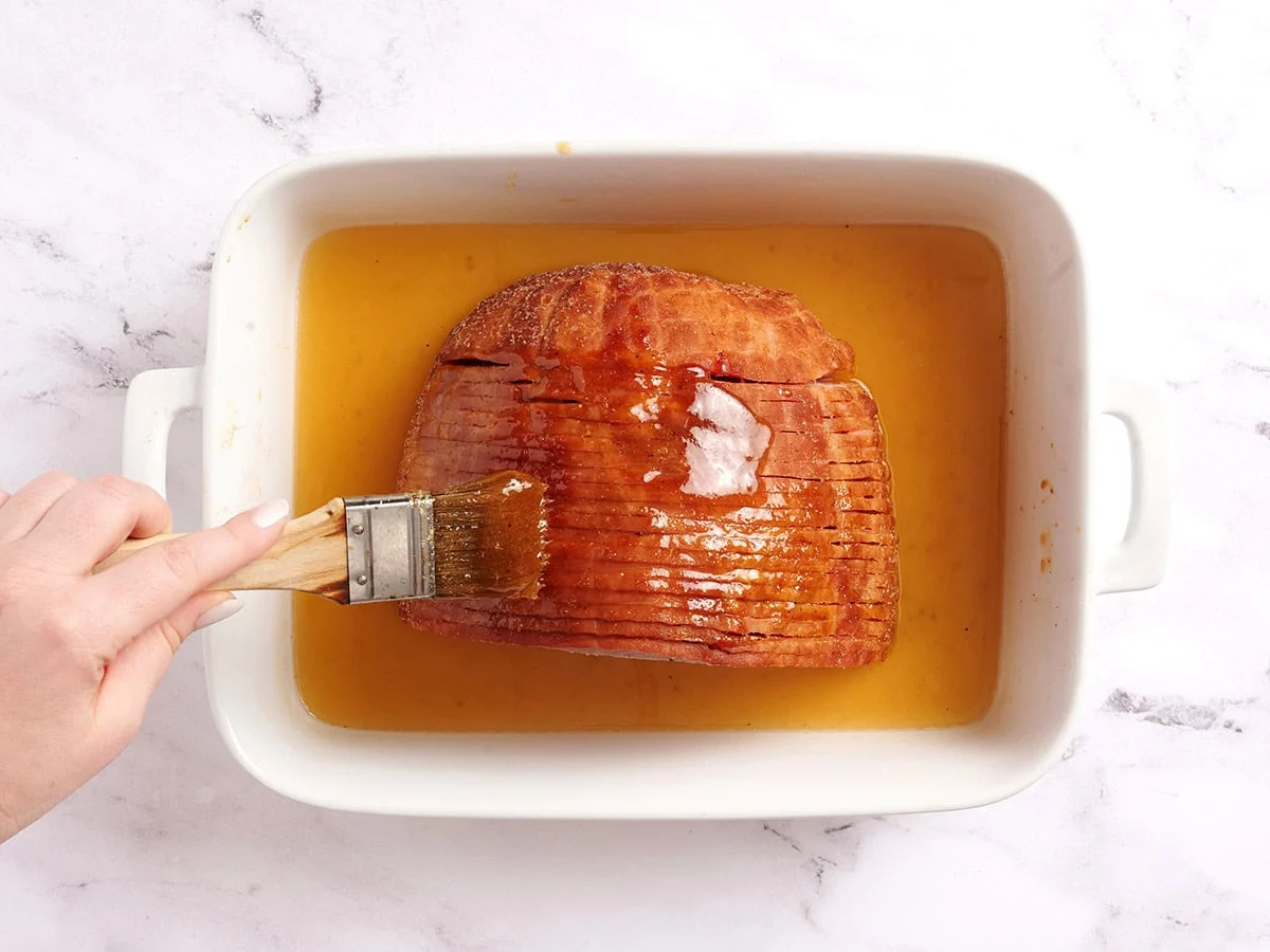 Un pincel rociando un jamón glaseado con miel a mitad de la cocción.