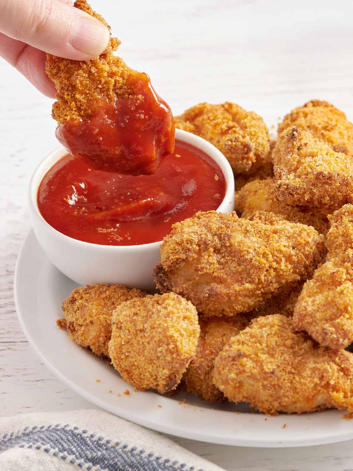 Vista lateral de un plato de nuggets de pollo caseros con una mano sumergiendo una pepita en una olla de salsa de tomate.