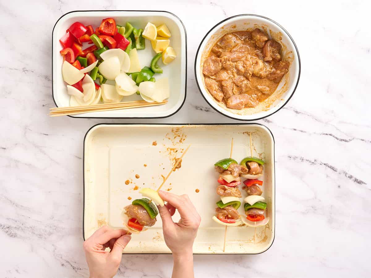 Una mano ensartando pimiento morrón, cebolla y pollo en una brocheta.