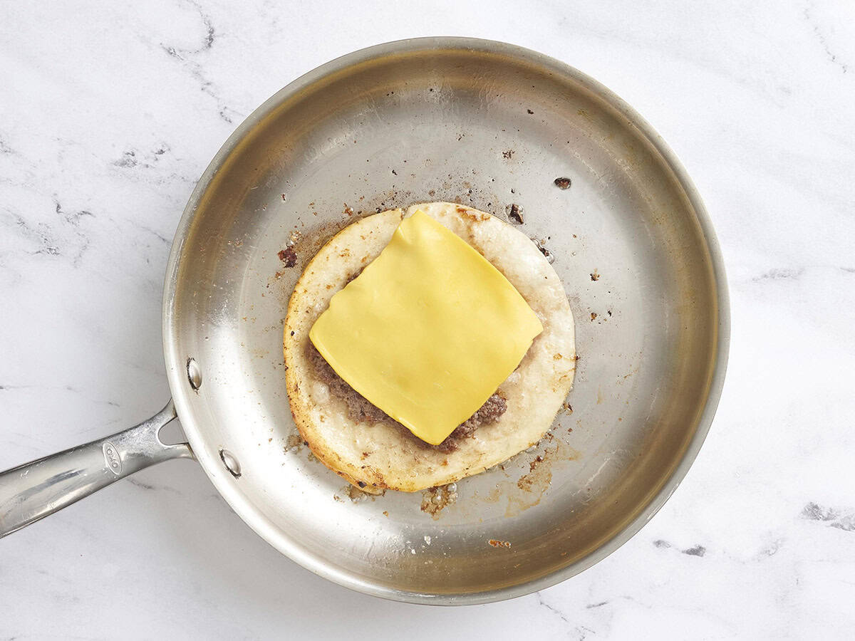 Un taco de hamburguesa aplastado en una sartén cubierto con una rebanada de queso americano.