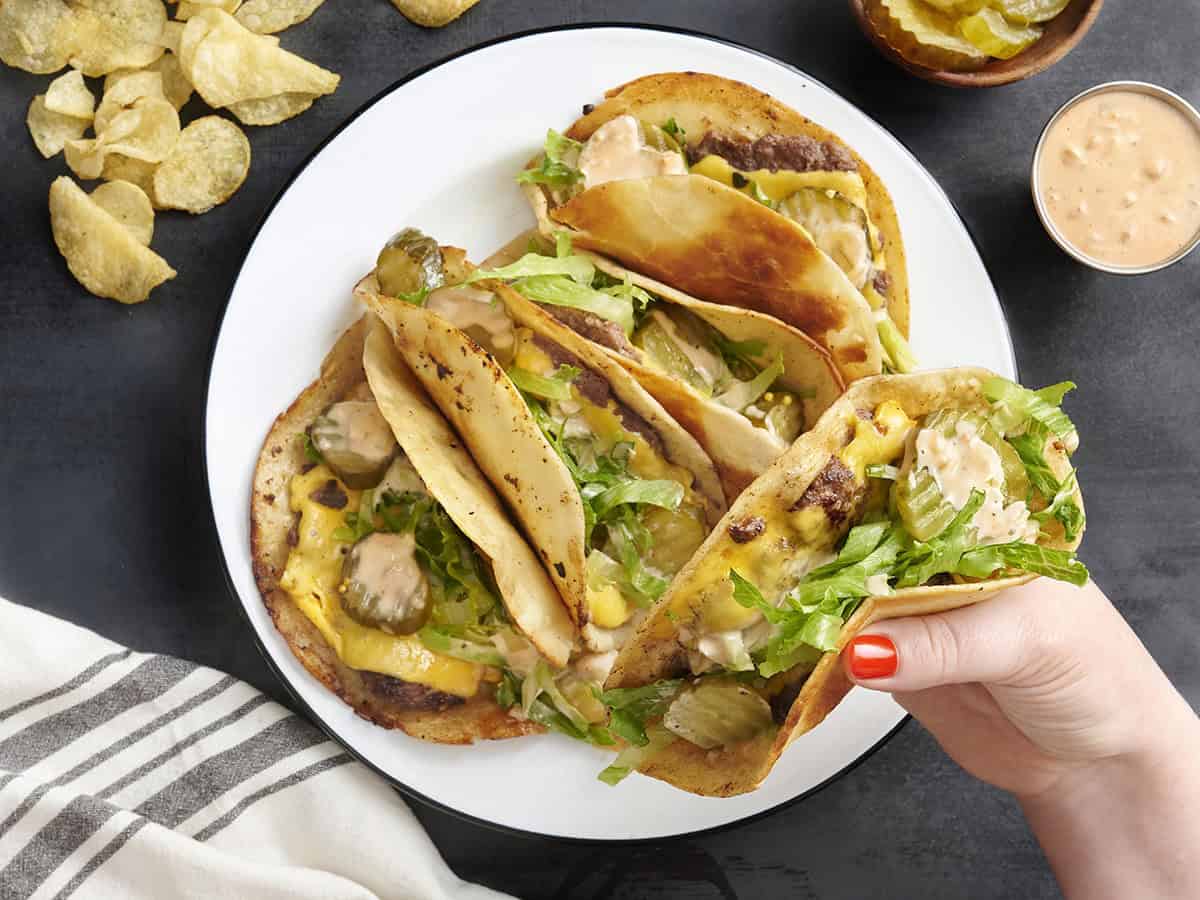 Vista aérea de un plato de tacos de hamburguesa caseros con una mano sosteniendo uno.