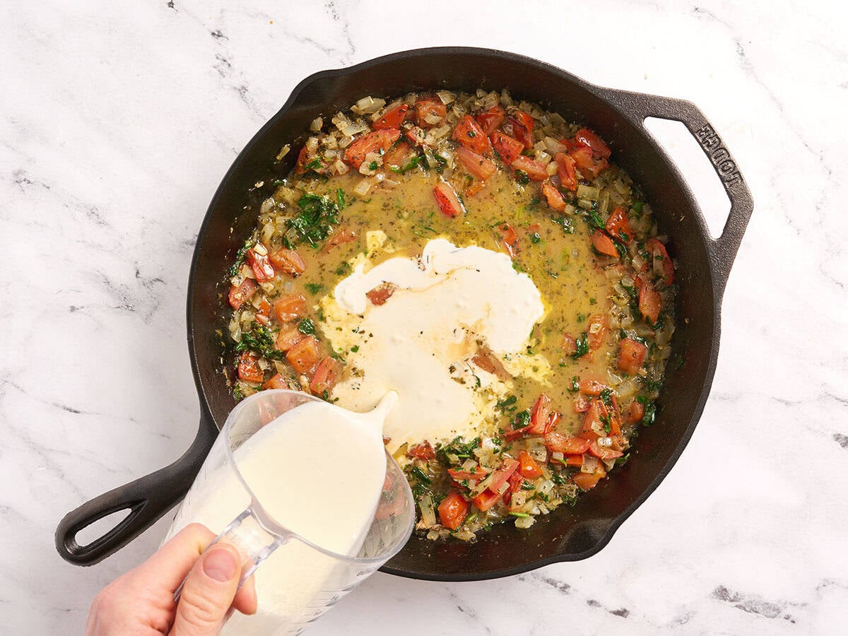 Crema espesa agregada a una sartén con cebollas, tomates y espinacas.