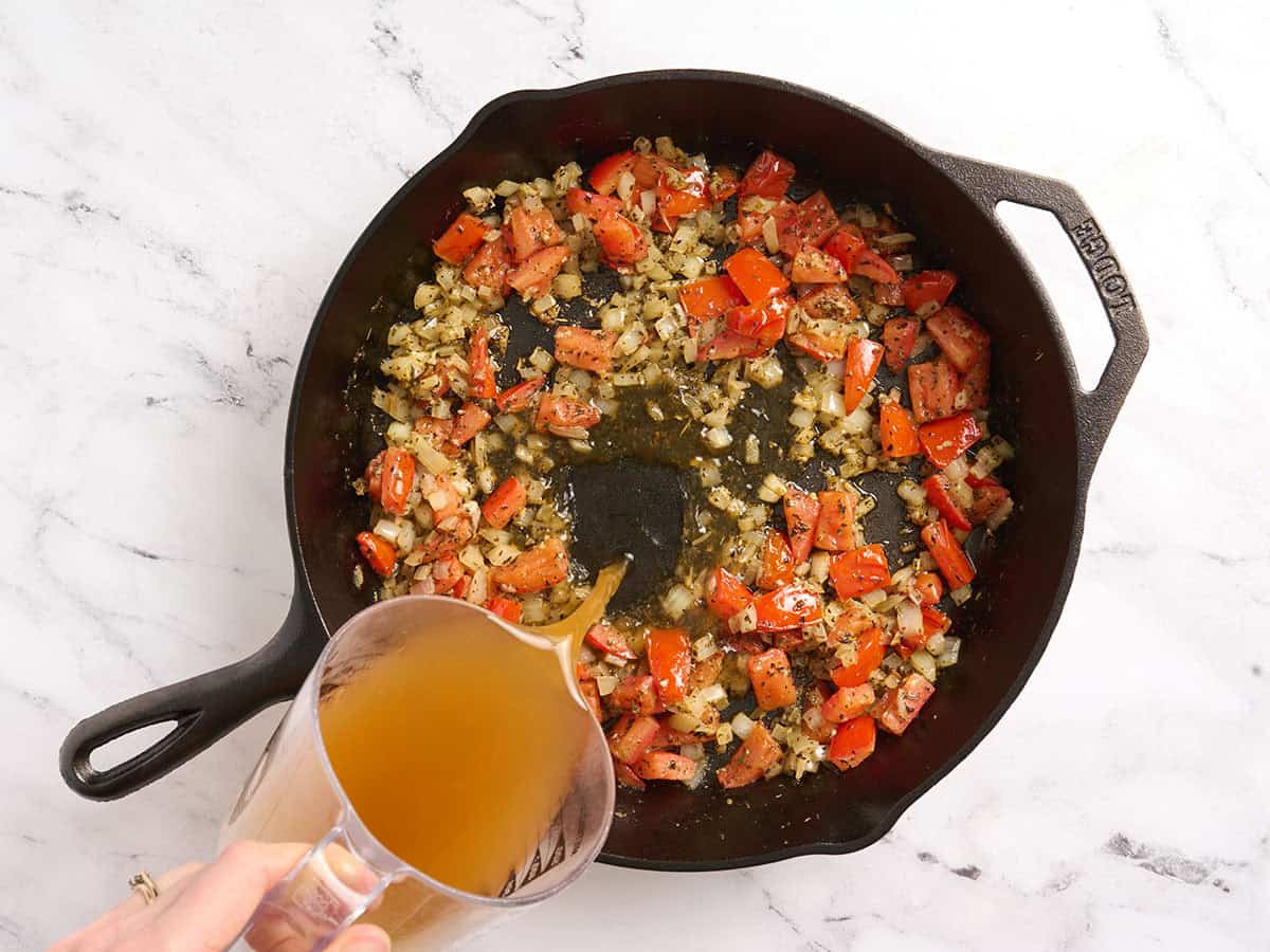 Caldo de verduras añadido a una sartén con cebollas y tomates.