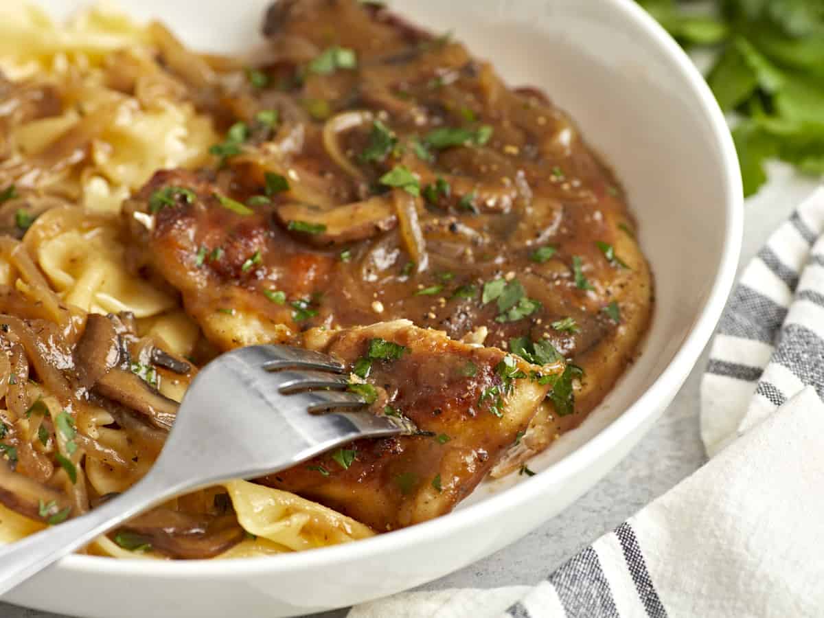 un tenedor que sostiene un trozo de pollo marsala sobre un plato poco profundo.