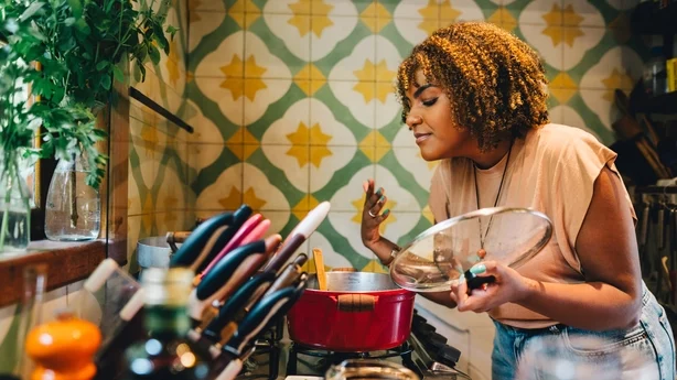 Mujer sonriendo con los ojos cerrados mientras huele comida cocinando en una olla en una estufa en su cocina en casa