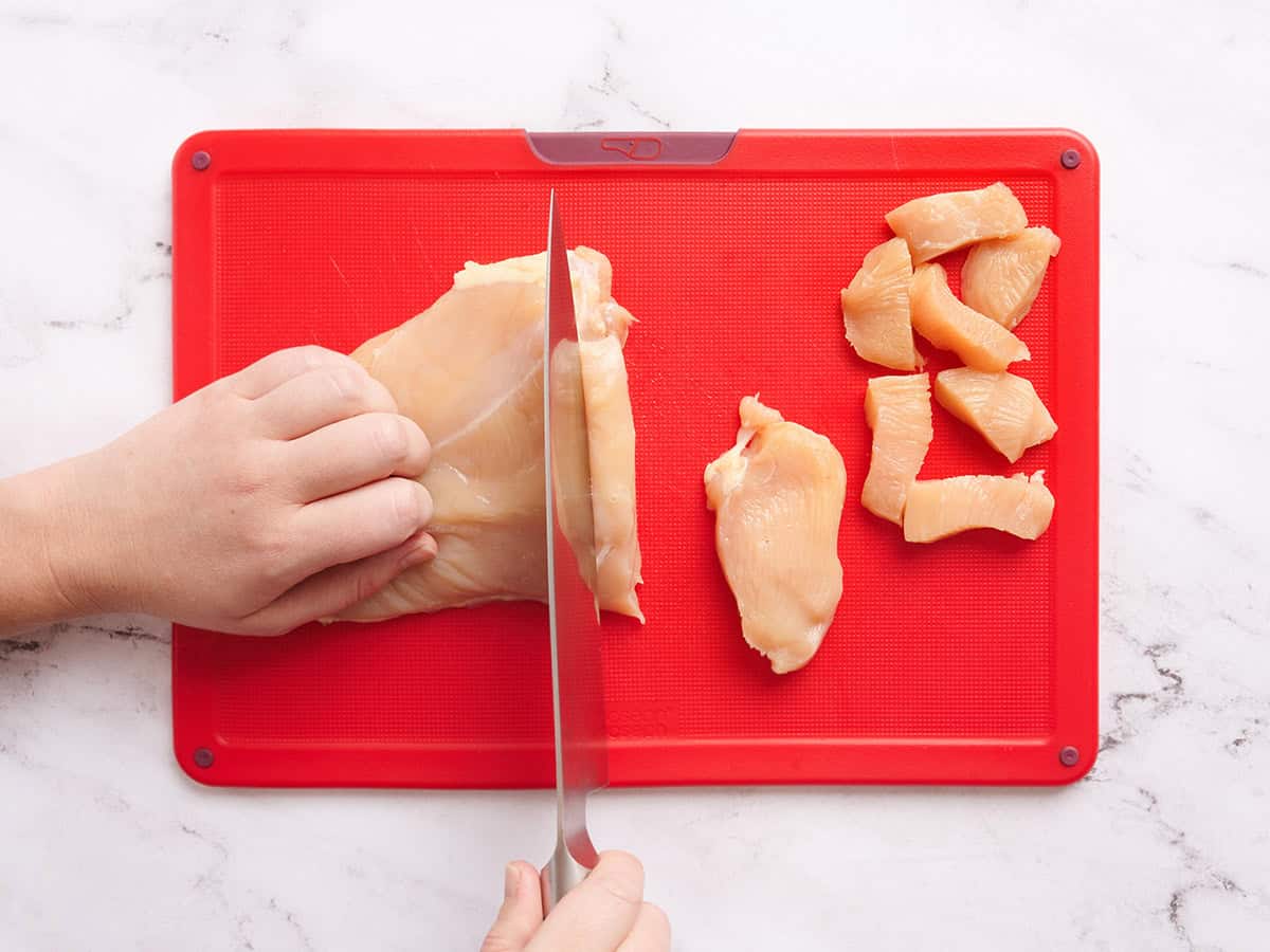 Manos usando un cuchillo para cortar una pechuga de pollo en trozos.