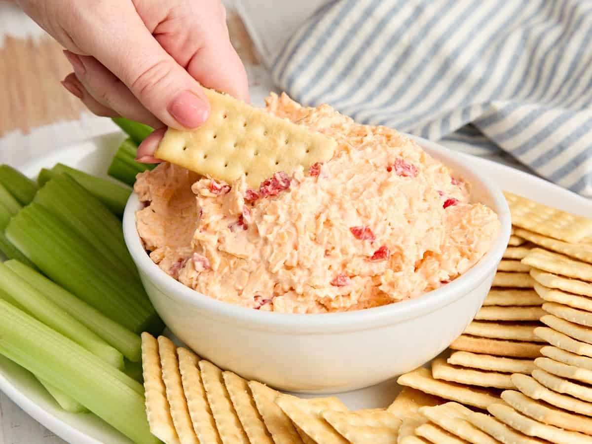 Vista lateral de un plato de salsa de queso pimiento sureño con una mano sumergiendo una galleta.