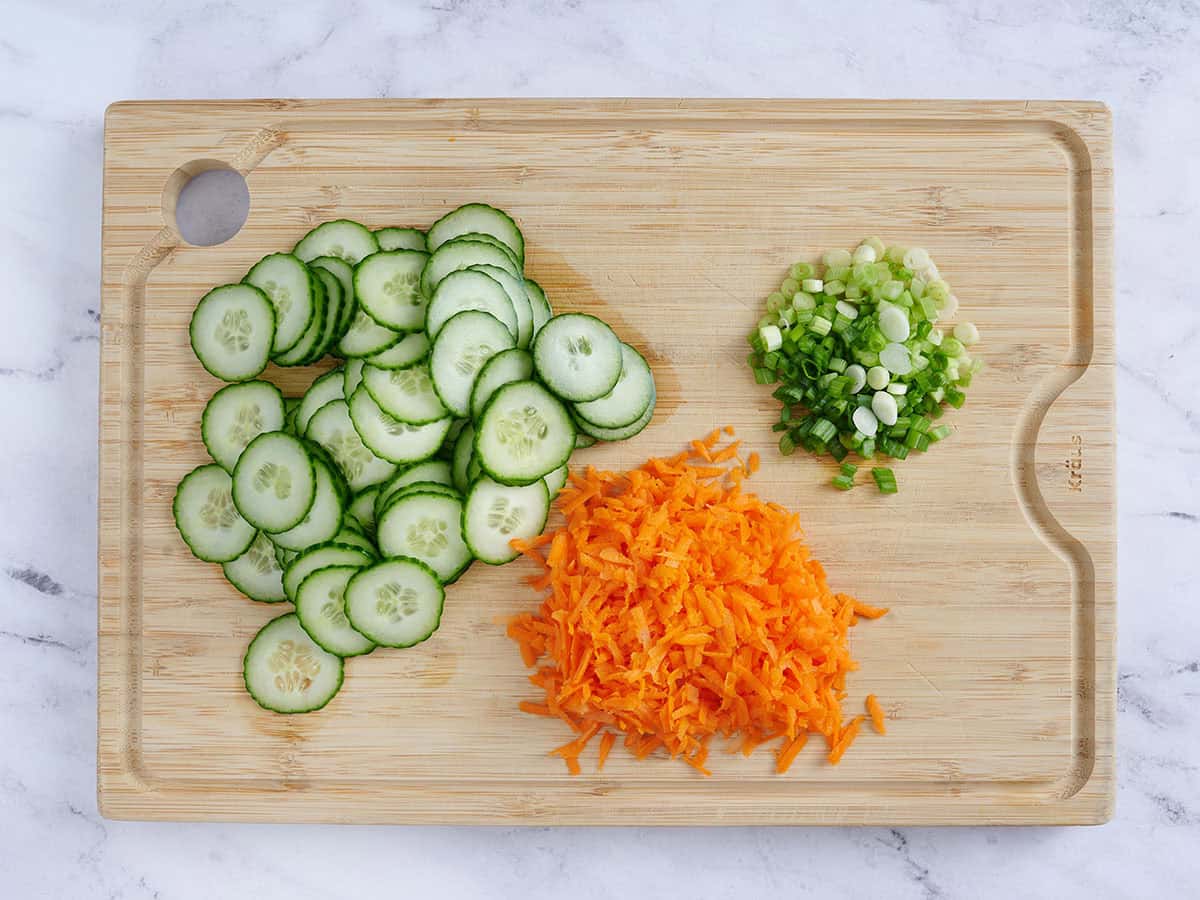 Pepino en rodajas, zanahorias ralladas y cebollas verdes picadas en una tabla de cortar de madera.