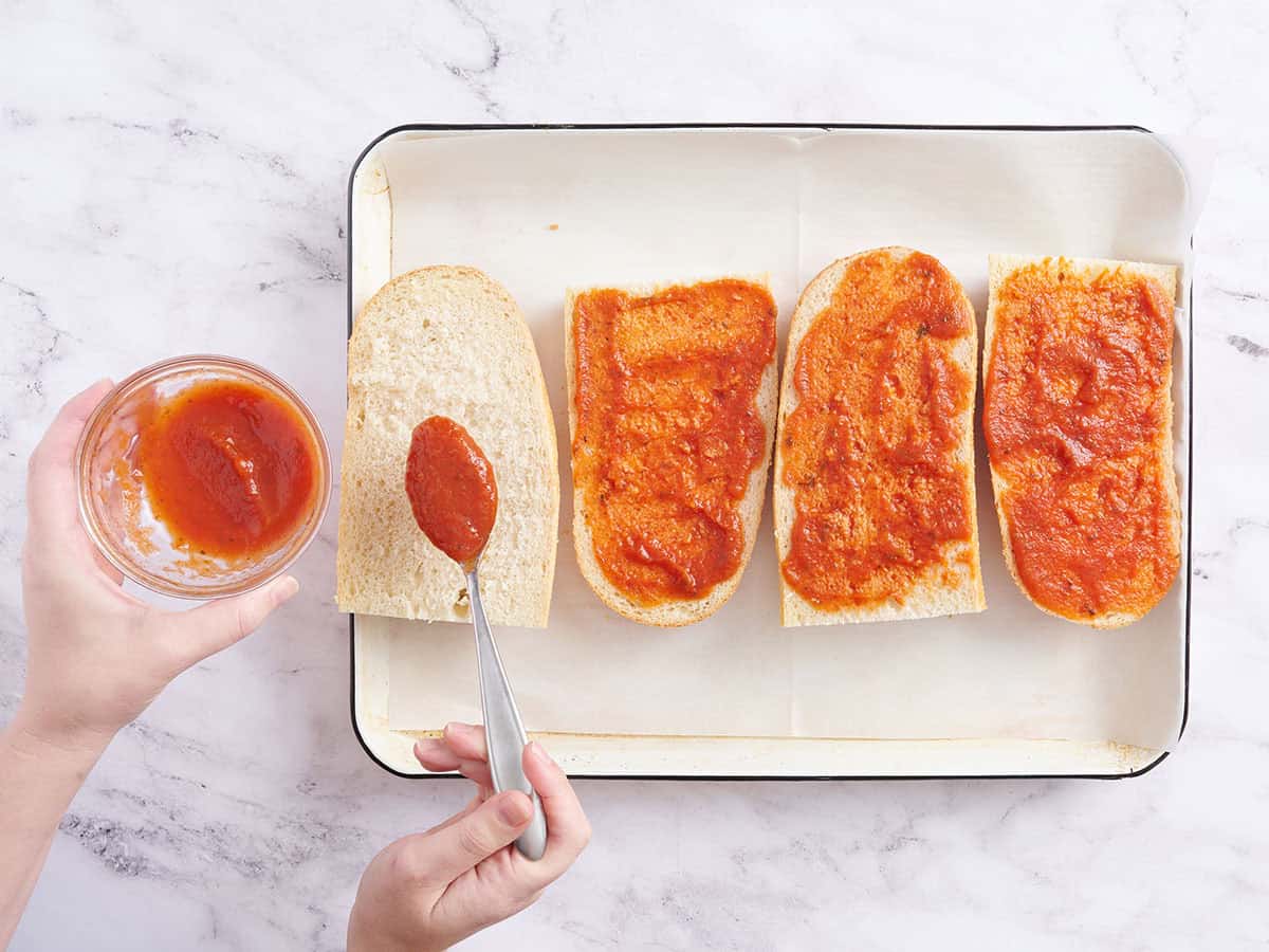 Una mano echando salsa para pizza sobre cuartos de pan francés.