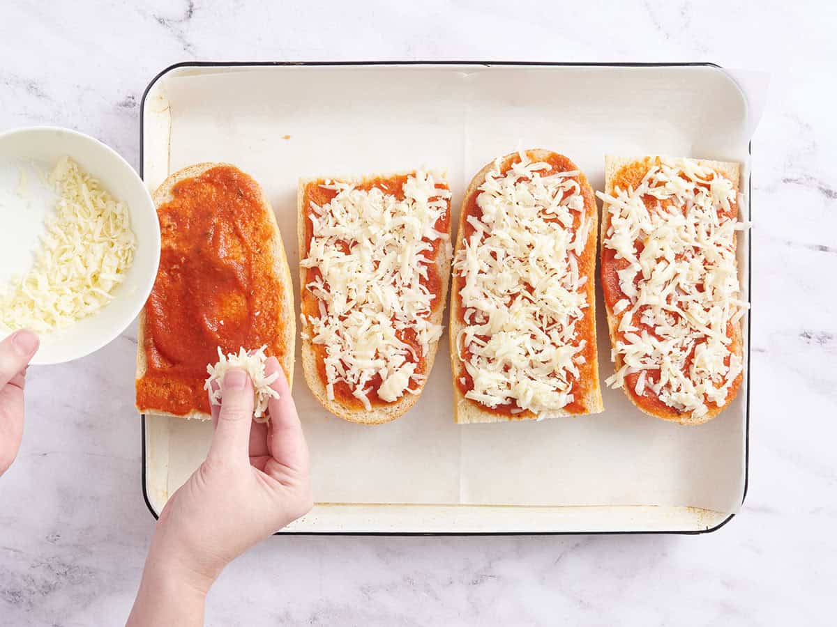 Una mano espolvoreando queso rallado sobre pizzas de pan francés.