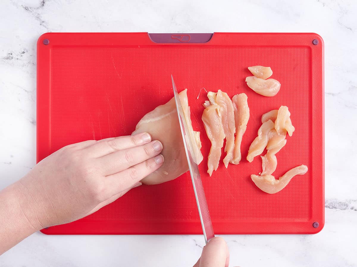 Manos cortando pechuga de pollo en una tabla para cortar.
