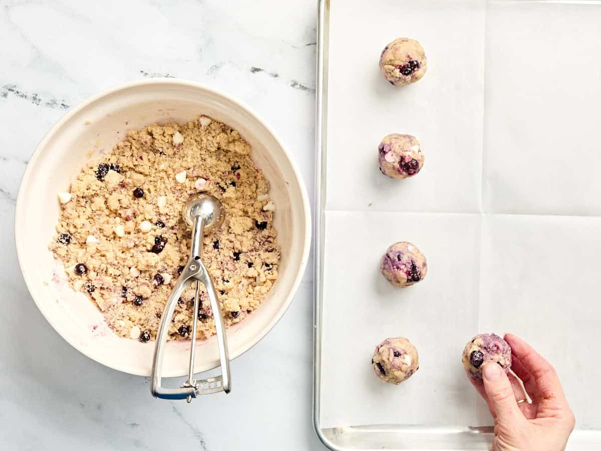 Un bol de masa para galletas de arándanos en un bol con una cuchara, junto a las bolas de masa para galletas en una bandeja para hornear.