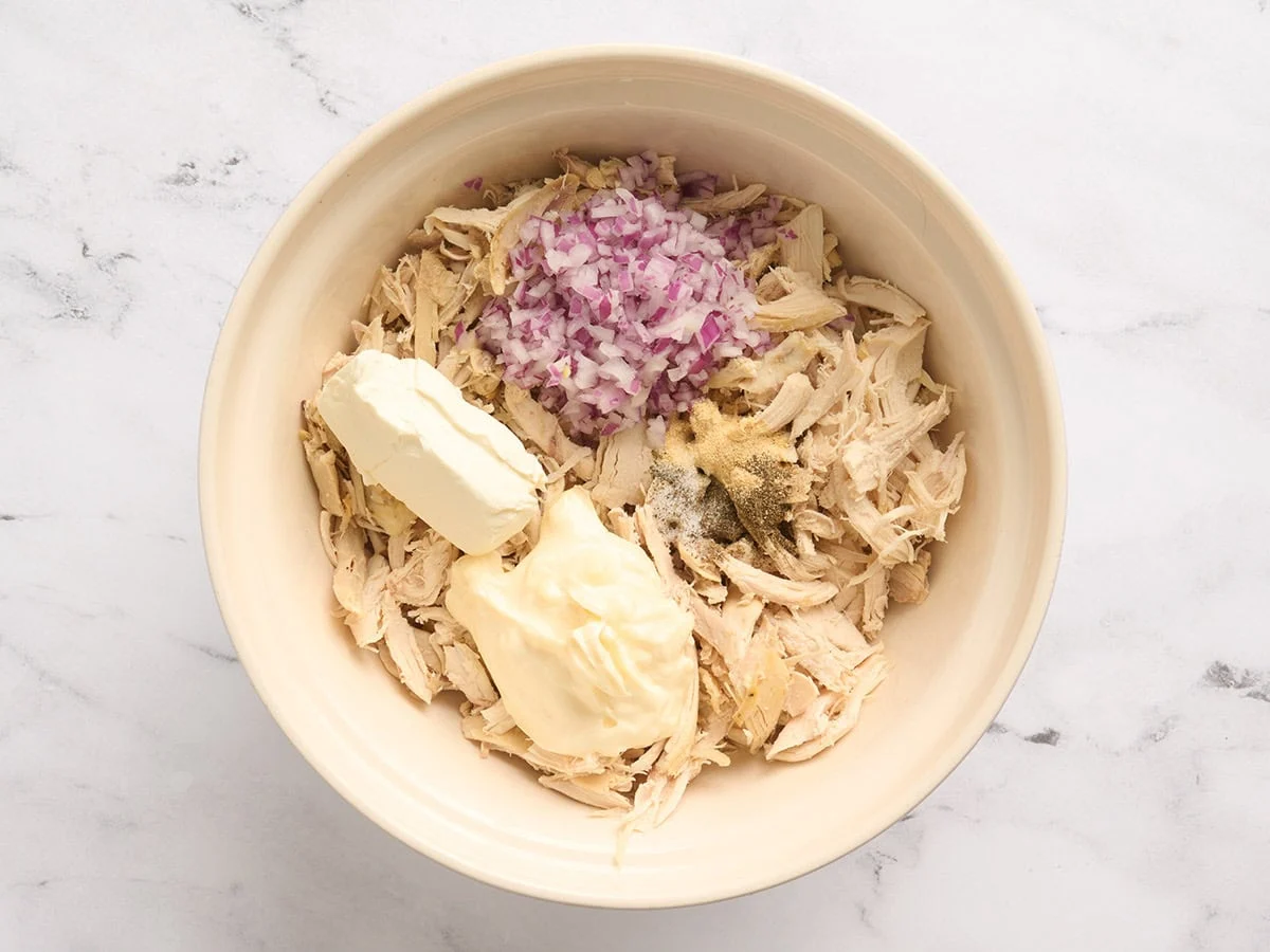 Pollo desmenuzado, cebolla morada picada, mayonesa, queso crema y condimentos en un tazón.