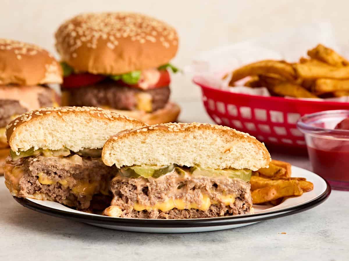Vista lateral de una jugosa hamburguesa Lucy cortada por la mitad en un plato con más hamburguesas y una canasta de papas fritas al fondo.
