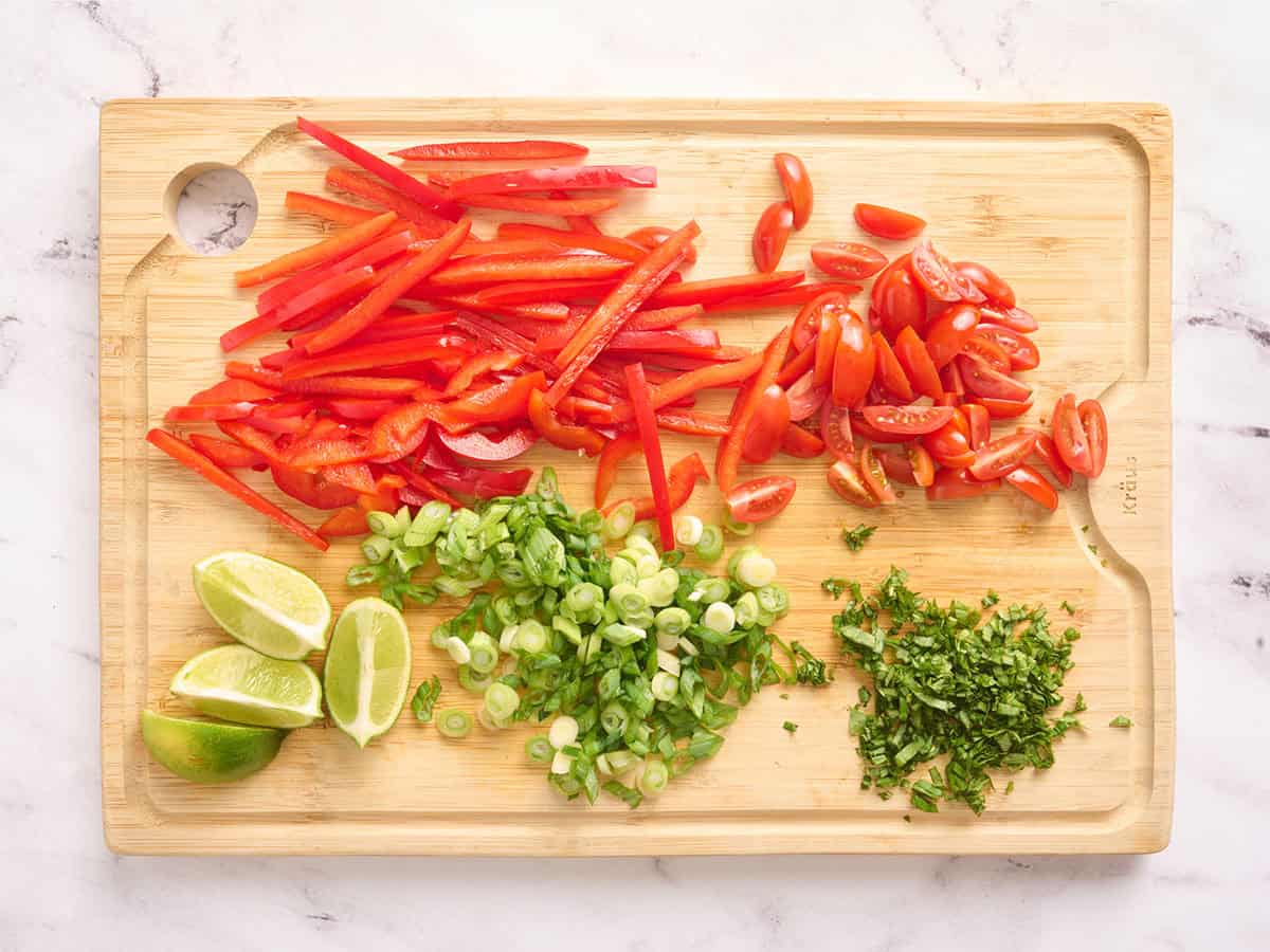 Pimiento rojo en rodajas, cebolla verde en rodajas, rodajas de limón en cuartos, tomates uva en rodajas y albahaca picada sobre una tabla de cortar de madera.