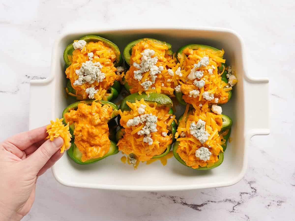 Mitades de pimiento verde rellenas de pollo búfalo, con una mano espolvoreando queso cheddar y queso azul encima.