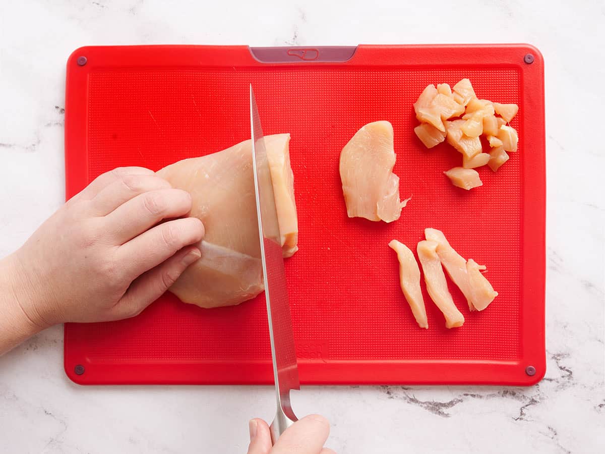 Una mano usando un cuchillo para cortar la pechuga de pollo en trozos.