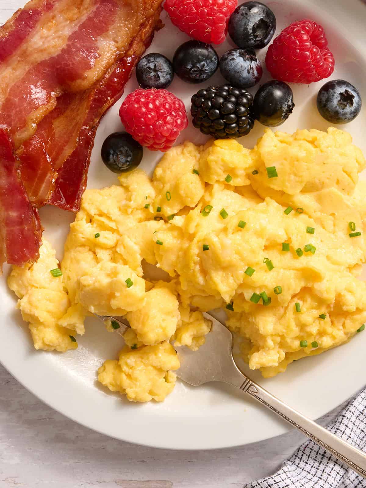 Vista aérea de huevos con requesón en un plato con tocino y ensalada de frutas.