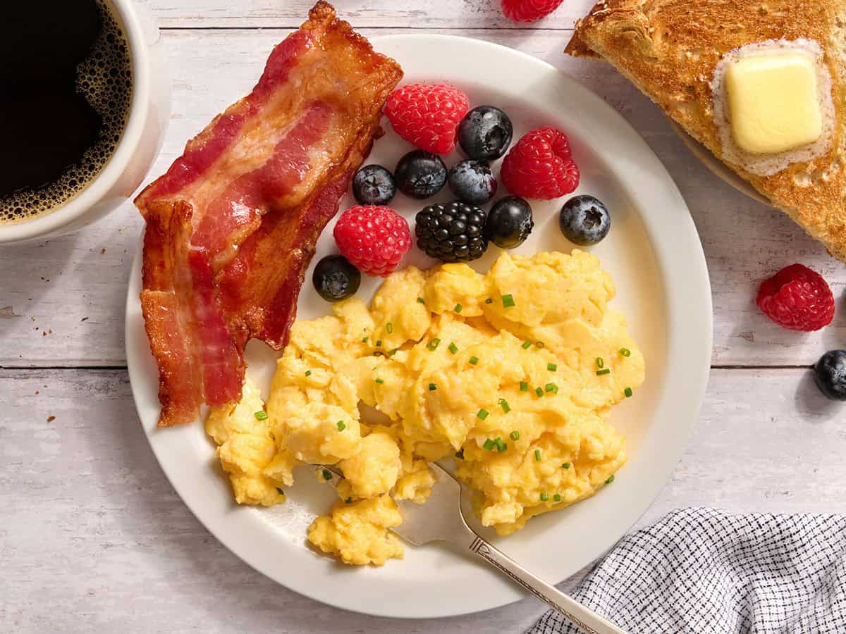 Vista aérea de huevos con requesón en un plato con tocino y ensalada de frutas.