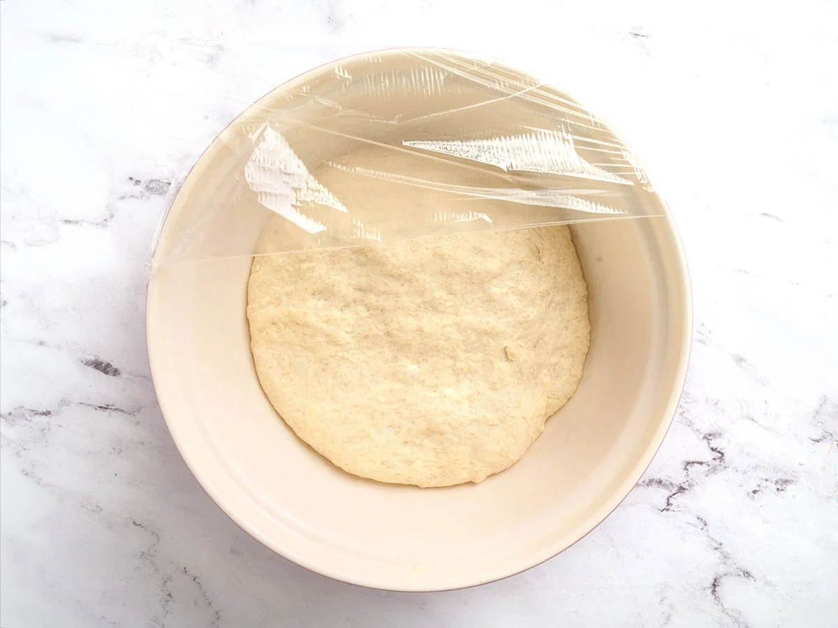 Masa de pan de pita en un bol después de fermentar y cubierta con film transparente.