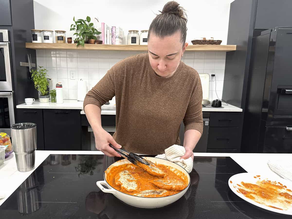 Una mujer cocinando pollo cremoso con tomate en una sartén.