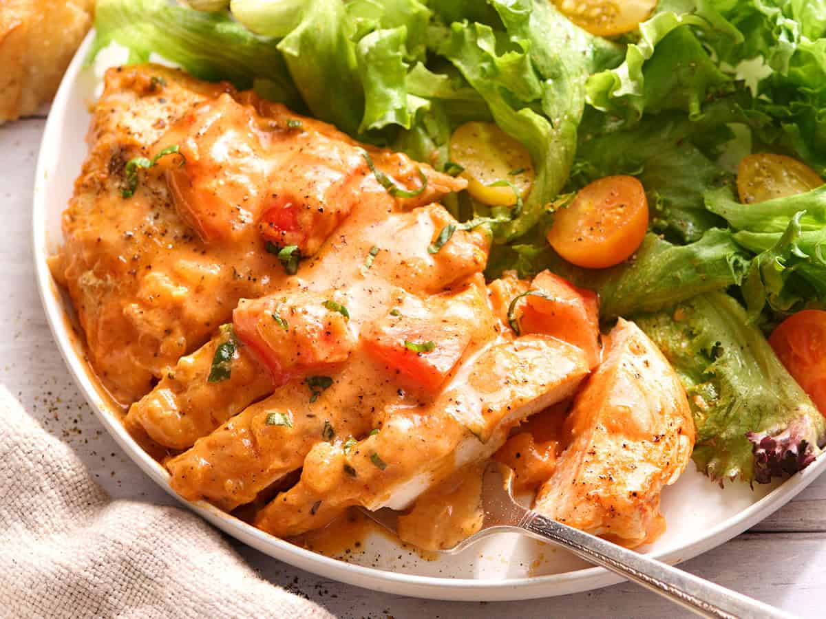 Vista lateral de una pechuga de pollo cremosa con tomate cortada en rodajas en un plato con ensalada verde.