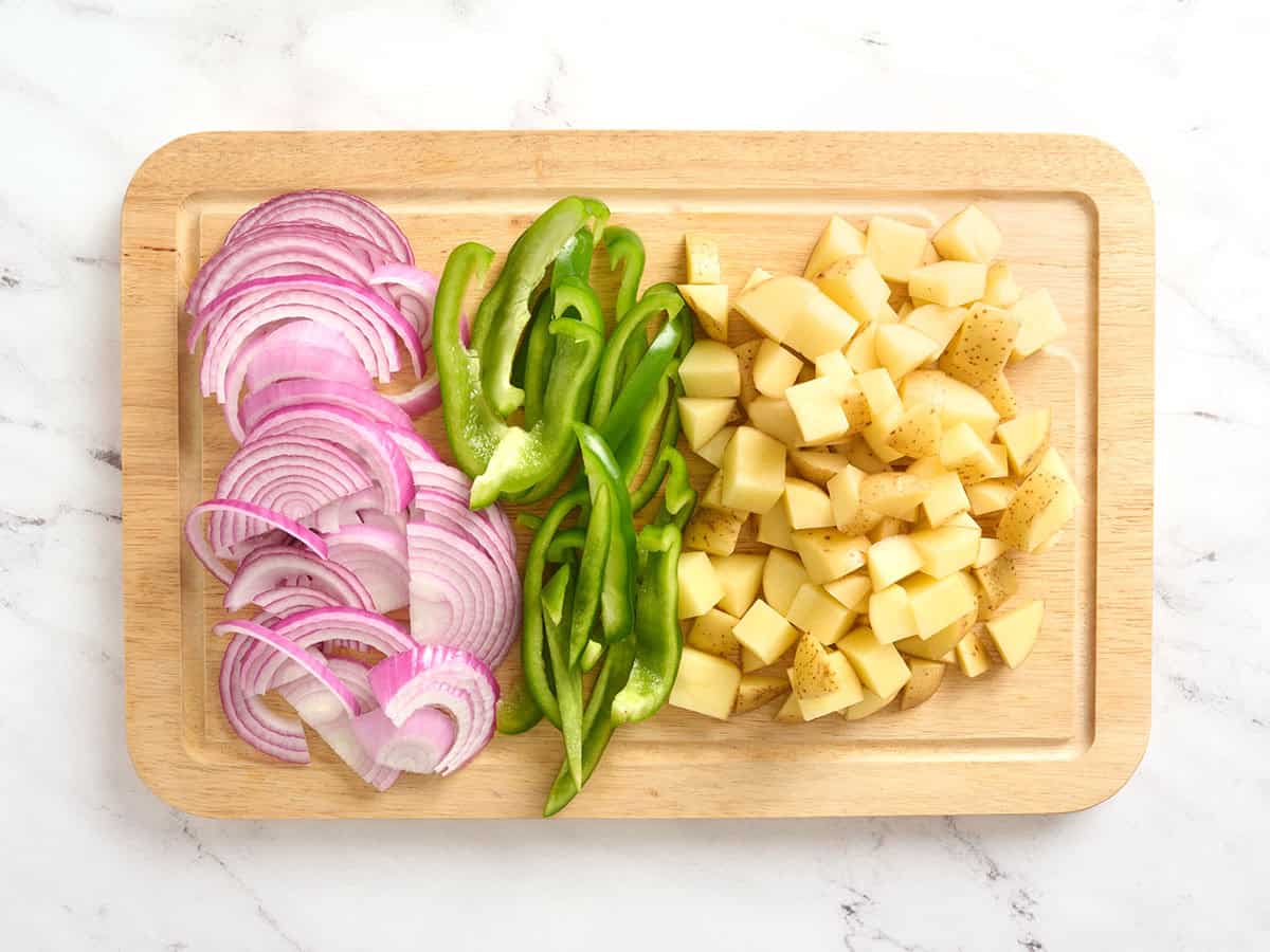 Cebollas rojas en rodajas, pimientos verdes y patatas cortadas en cubitos sobre una tabla de cortar.