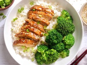 Overhead view of sliced teriyaki chicken on a bed on rice on a plate with broccoli.