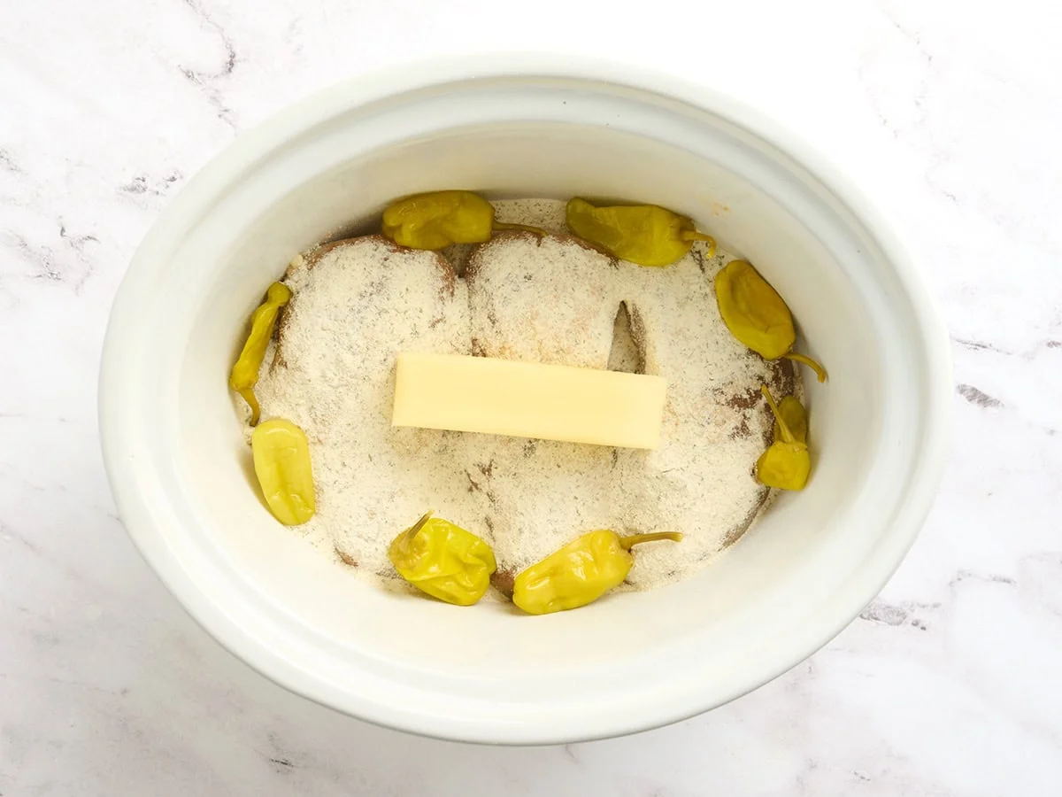 Pechugas de pollo en una olla con condimentos, una barra de mantequilla y pimientos pepperoncini para hacer pollo Mississippi.