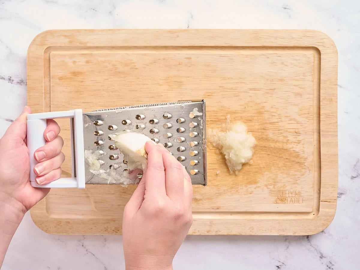 Manos usando un rallador de queso para rallar una cebolla blanca.