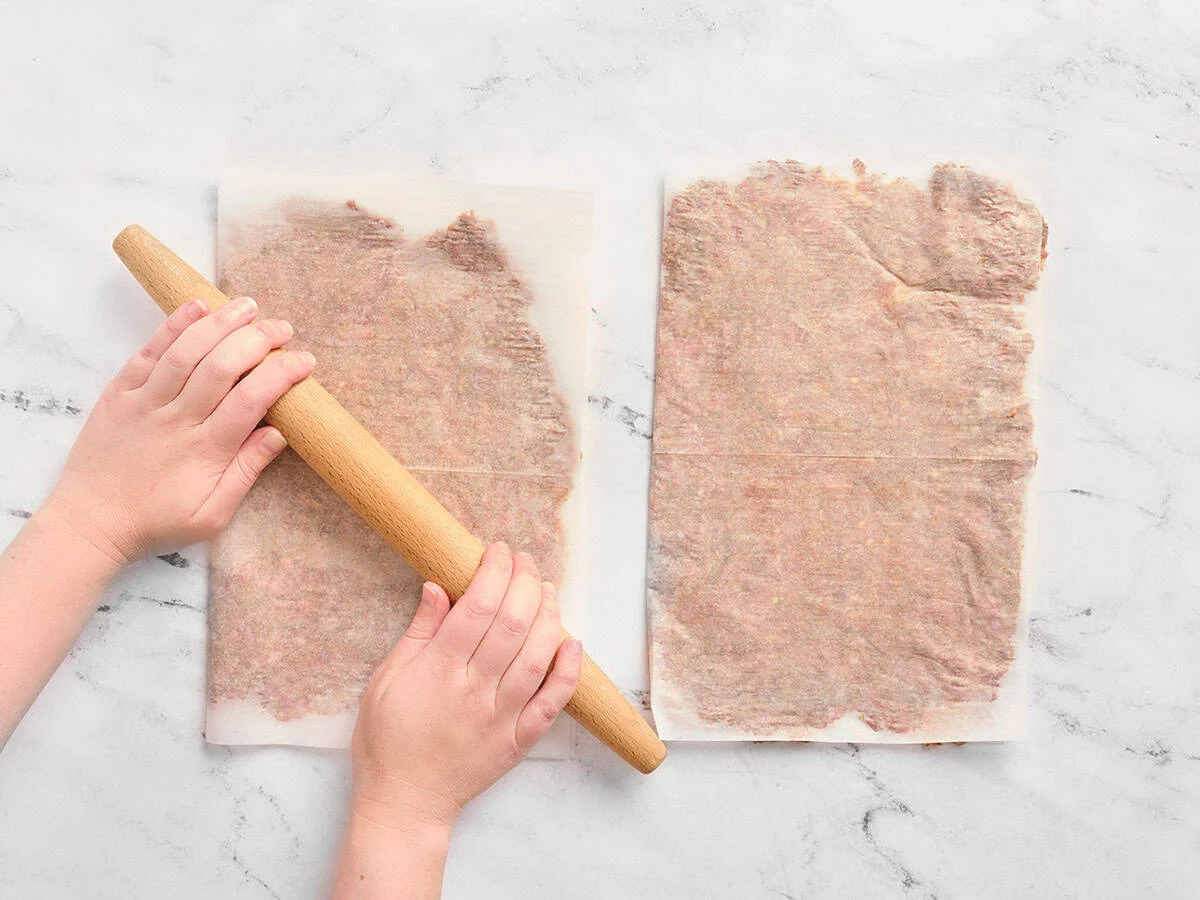 Manos usando un rodillo para aplanar la carne molida entre dos trozos de papel pergamino hasta que quede aplanada.